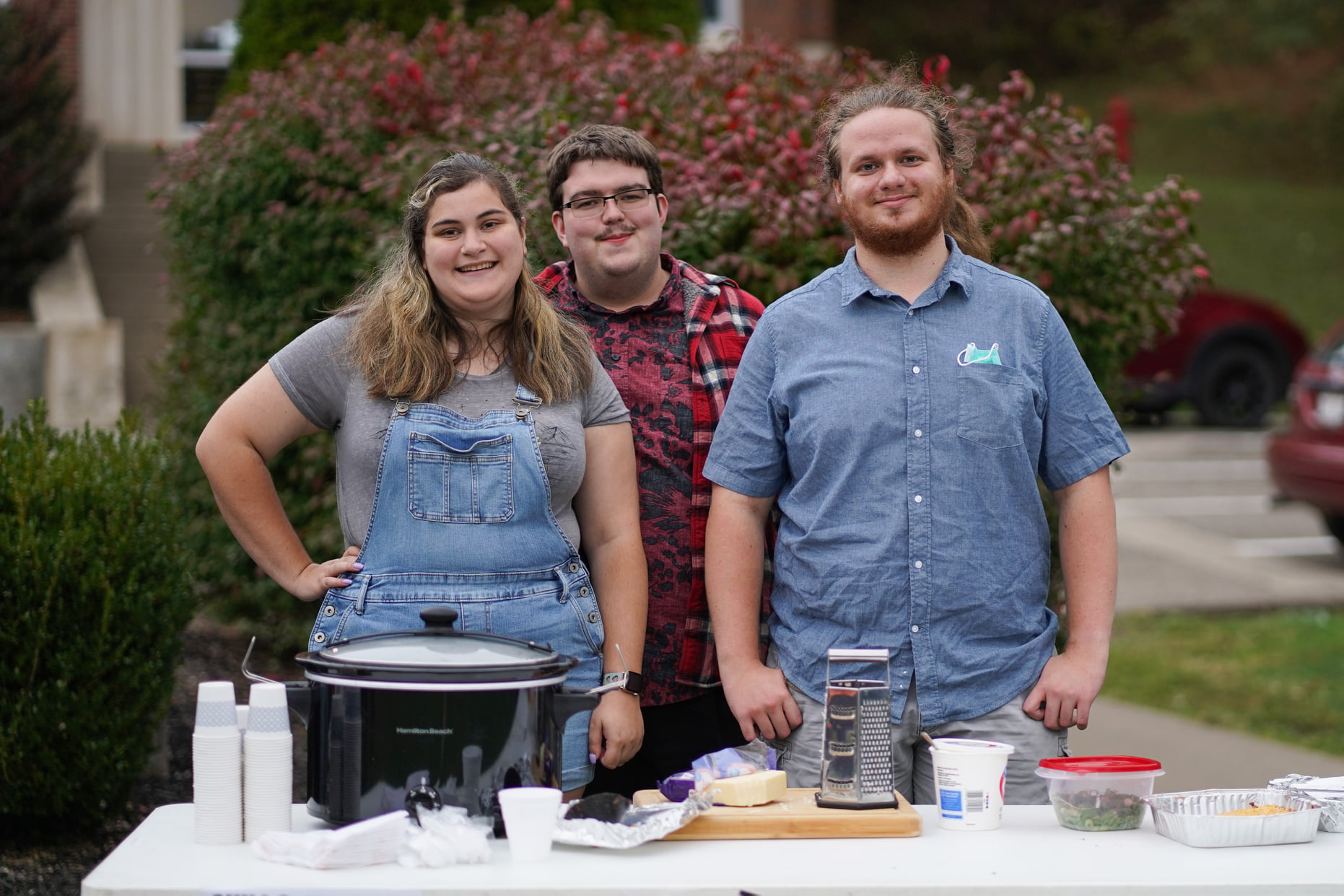 Three people standing behind an outdoor table with a Crock-Pot of chili and toppings
